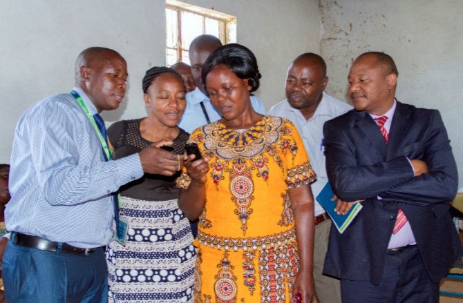 A member of one of the Chama Solutions Group (pressing a phone) demonstrating how they conduct group business through the Chama Solutions Digital Platform. Looking on are officials from Nuru Kenya and KCB