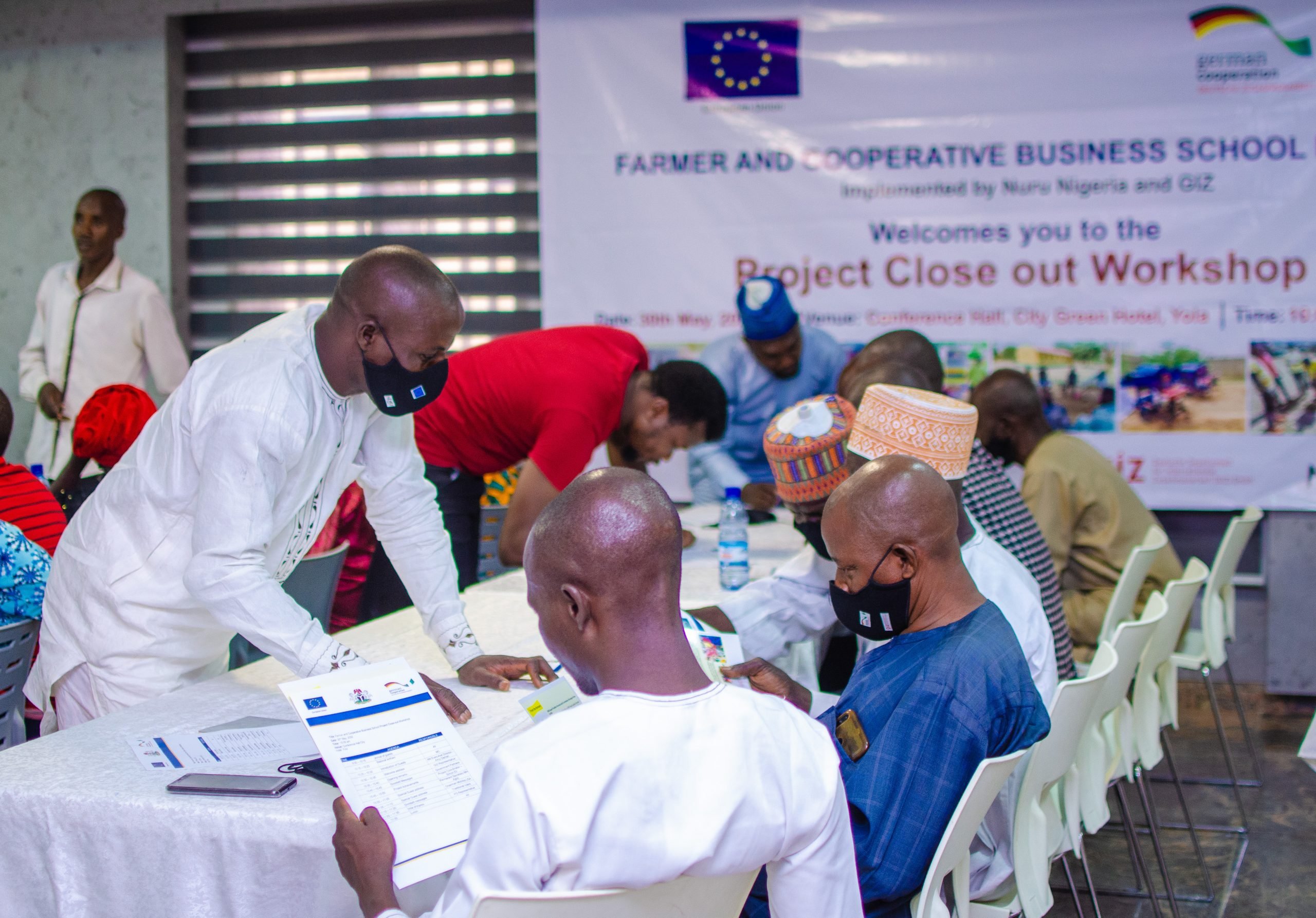 Group of men, primarily in traditional Nigerian dress, sit and stand around a table and look at event materials. Sign in the background reads "Farmer and Cooperative Business School Project, Implemented by Nuru Nigeria and GIZ, Welcomes you to the Project Close out Workshop"