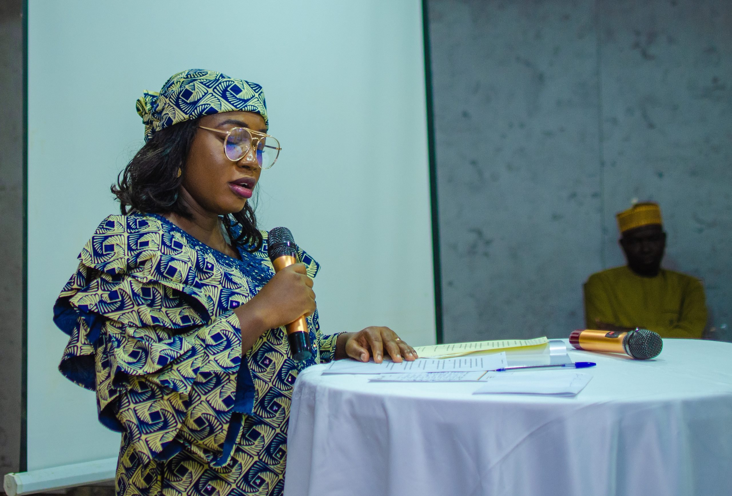 Woman in traditional Nigerian dress stands in front of a linen covered podium, speaking into microphone