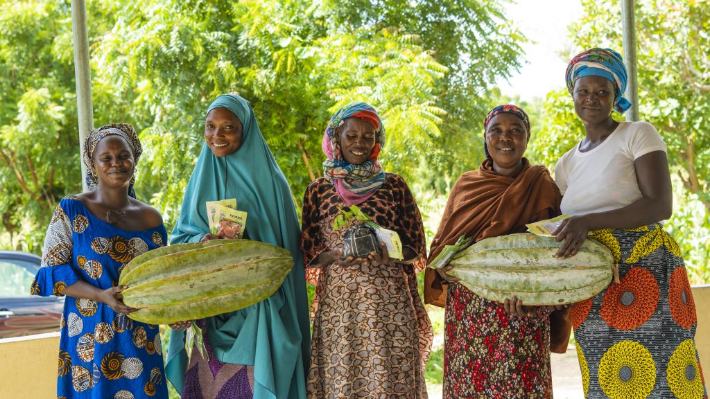 women farmers stand holding fluted pumpkins and smiling at the camera