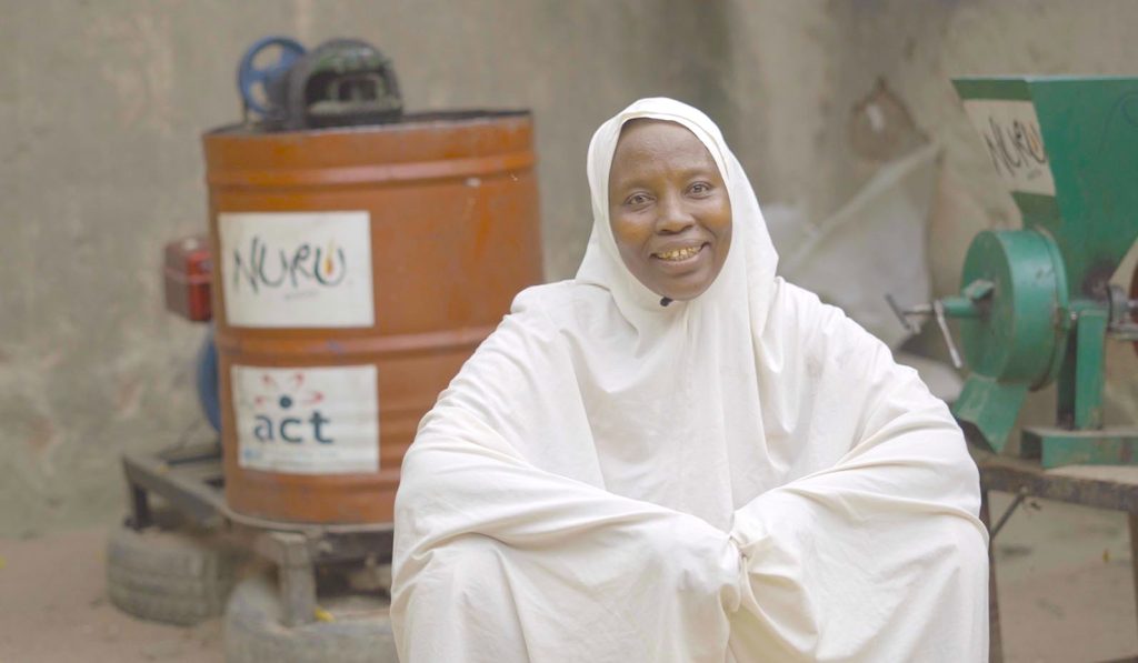 woman sits outside in front of groundnut processing equipment and smiles at the camera