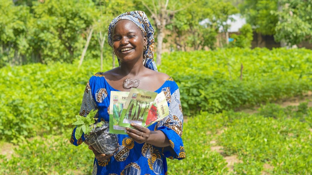 woman smiling in a field in Nigeria while holding a plant and a packet of seeds